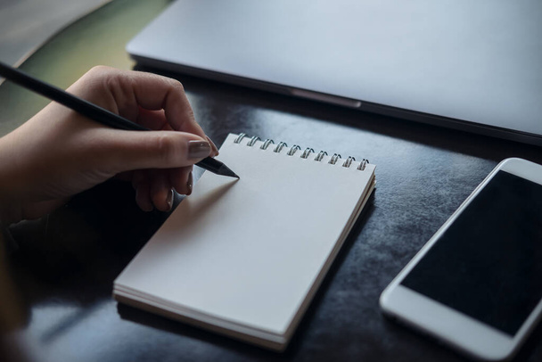 Крупним планом зображення рук жінки, що пишуть на білому порожньому блокноті з мобільним телефоном і ноутбуком на столі
  - Фото, зображення