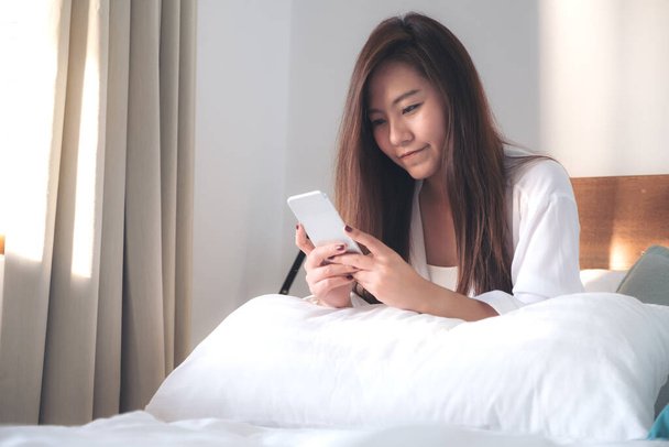 Güzel bir Asyalı kadının portre resmi yatakta oturmuş, mutlu bir şekilde uyandıktan sonra cep telefonuna bakıyor ve kullanıyor. - Fotoğraf, Görsel