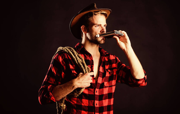 Sheriff concept. Brutal cowboy drinking alcohol. Western culture. Western life. Man wearing hat hold rope and flask. Lasso tool of American cowboy. Man handsome unshaven cowboy black background - Фото, зображення