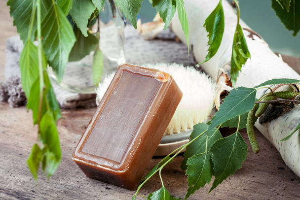 Il sapone marrone scuro con l'aggiunta di catrame di betulla si trova su una spazzola da massaggio. Rami di betulla freschi, fondo in legno. Cosmetici naturali - Foto, immagini