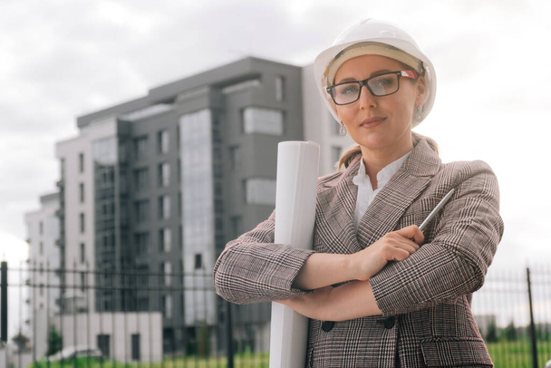 Stylish woman with glasses in uniform in a white helmet against the background of the business building. In the hands of documents with the drawings of the building. - 写真・画像