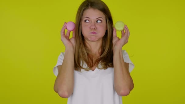 Woman looks at a pink, yellow macaroons in hands with pouting cheeks, mouth - Footage, Video