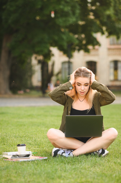Schockiertes Mädchen in legerer Kleidung sitzt mit Büchern und Laptop auf dem Rasen in der Nähe der Universität, blickt mit erstaunter Miene in den Bildschirm. Emotionale Schülerin schockiert durch Aufgaben, Lernen in der Pause. Freizeitstudent - Foto, Bild