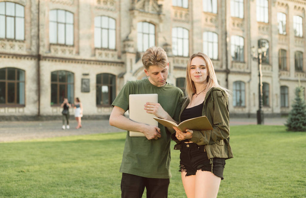 Young couple stands on the grass on a background of a college building with a laptop and a notebook, guy reads information from a notebook of girl with a serious face, lady looks in camera and smiles - Fotó, kép