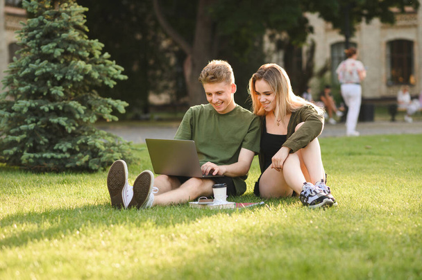 Couple of happy students using laptop while sitting on university campus lawn, looking at screen and smiling. Smiling girl and young man studying with laptop at park break. Student leisure. - Фото, изображение
