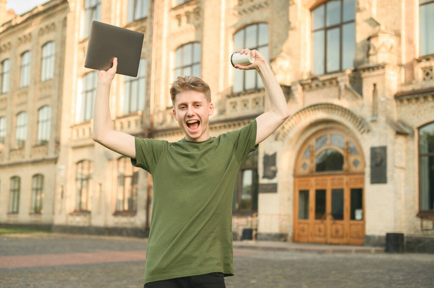 Portrét radostného mladíka v neformálních šatech na pozadí univerzitní budovy, zvedl ruce a s úsměvem se zadíval do kamery. Mladý student složil zkoušku, je šťastný. - Fotografie, Obrázek