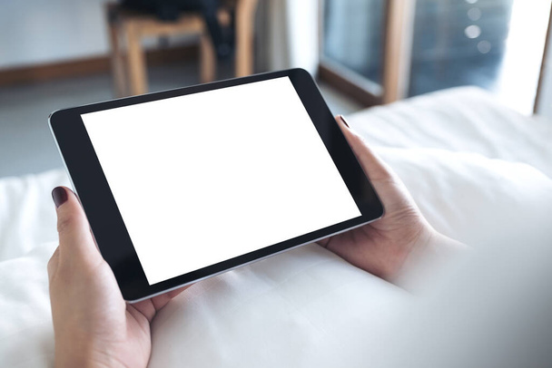 Mockup afbeelding van de handen van de vrouw houden zwarte tablet pc met blanco desktop wit scherm terwijl zitten op een wit bed - Foto, afbeelding