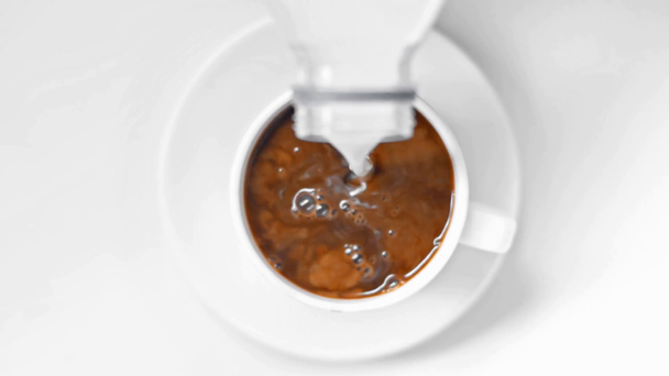 górny widok mleka wylewanie z butelki w filiżance czarnej kawy na białym - Materiał filmowy, wideo