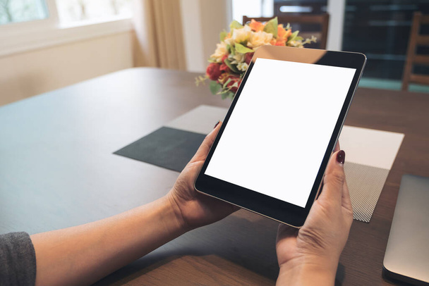 Evdeki ahşap masanın üzerinde boş masaüstü beyaz ekranı olan siyah tableti tutan ellerin görüntüsü - Fotoğraf, Görsel