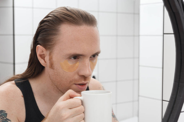 Нездоланний чоловік з плямами під очима і чашкою кави дивиться на себе у дзеркало ванної кімнати
. - Фото, зображення