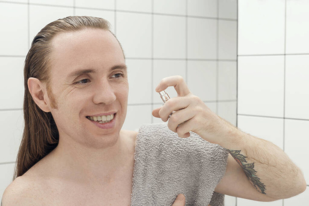 Un homme avec des tatouages et des pulvérisations de cheveux longs lotion après-rasage dans la salle de bain - Photo, image