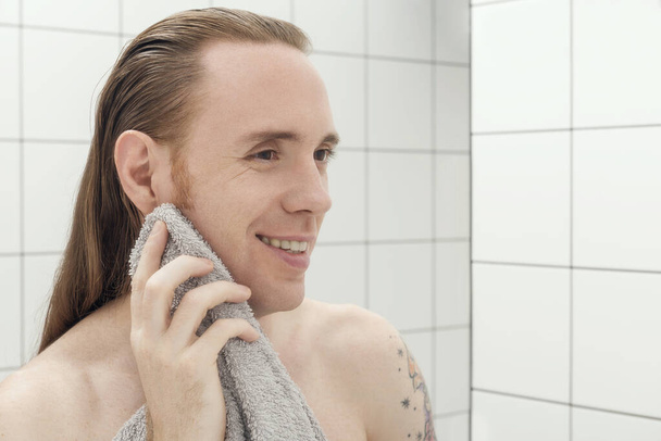 Ο άνθρωπος σκουπίζει το πρόσωπό του με μια πετσέτα και χαμογελά στην αντανάκλασή του στον καθρέφτη του μπάνιου. - Φωτογραφία, εικόνα