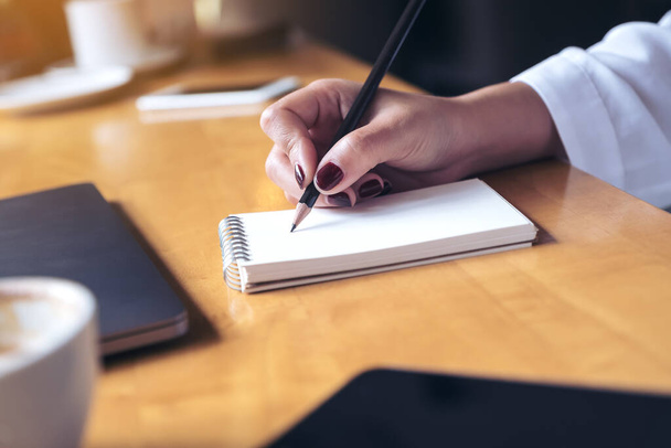 Imagen de primer plano de la escritura de la mano de la mujer en un cuaderno en blanco con computadora portátil, tableta y taza de café sobre fondo de mesa de madera
 - Foto, Imagen