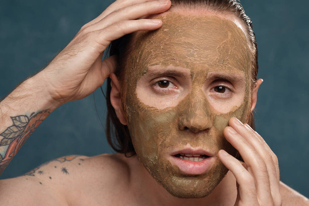 Posing man with wet long hair in green clay mask on his face waiting of the spa procedures. - Foto, afbeelding