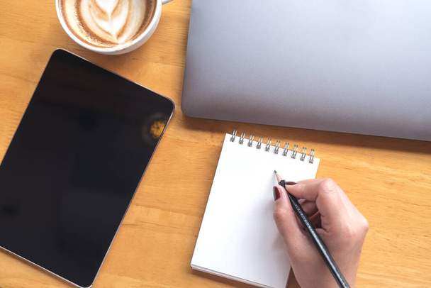 Widok z góry obraz dłoni kobiety trzymającej ołówek do pisania na pustym notebooku z tabletem, laptopem i kawą na drewnianym stole w biurze - Zdjęcie, obraz