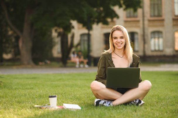 Happy girl sitting on lawn on background of university building with laptop on lap and books, looking into camera and smiling. Smiling lady student with laptop relaxing on lawn. - Photo, Image