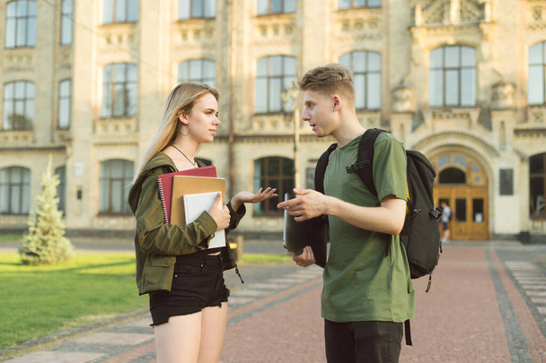 Два красивых студента, парень и девушка, разговаривают на фоне университетского здания, держа в руках книги, тетради и ноутбук. Пара молодых людей, болтающих в кампусе
. - Фото, изображение