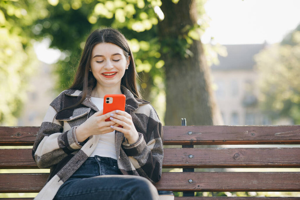 Young woman in trendy casual outfit sits on a bench in the city park, actively texts messages via her phone. Being online, modern technologies. - Фото, изображение