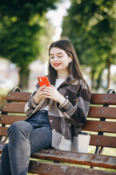 Girl wearing earphones and using smartphone listening music. Attractive woman browsing on mobile phone in park. City, urban background. - Photo, Image