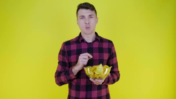 Mężczyzna w różowej koszuli w kratę je chipsy ziemniaczane ze szklaną miską na żółtym tle - Materiał filmowy, wideo