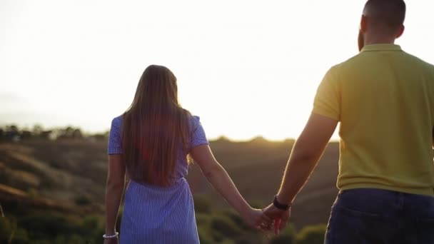 Rear view of loving young caucasian couple of beautiful girl and bearded guy holding hands in field on grass and enjoying wonderful evening sunset in slow motion. Romantic date and leisure in nature. - Footage, Video
