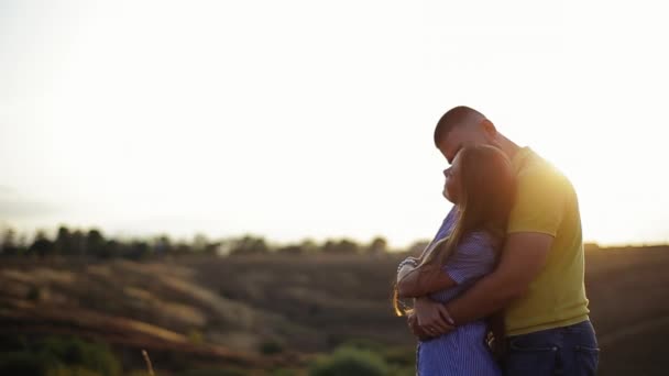 Tall young bearded guy in polo shirt hugs behind pretty girl in striped dress and hold hands in field against defocused background of sunset in evening. Romantic date in nature outside couple in love - Footage, Video