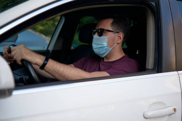 A man driving a car in a medical face mask during coronavirus outbreak, a taxi driver in a mask, protection from the virus. Driver in white car, hand on the steering wheel, looking out the open window. Quarantine, covid-19. - Photo, Image