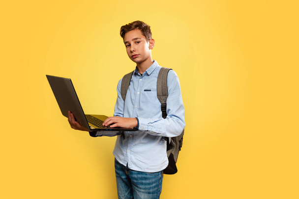 Concept of modern technologies and education. A young Caucasian boy in a blue shirt and backpack, holding an open laptop. Yellow background. Copy space - Photo, Image