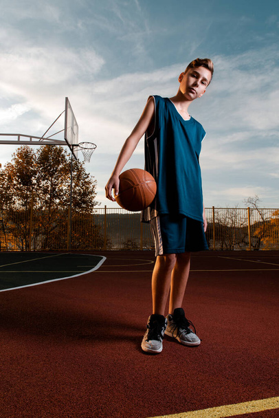 Deportes y baloncesto. Un joven adolescente en un chándal negro posa con una pelota de baloncesto en el patio de recreo. Vertical
 - Foto, imagen