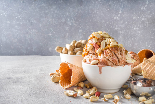 Peanut butter ice cream. Sweet peanut ice cream with peanut butter topping and a lot of peanuts, on grey stone background with icecream waffle cones - Foto, imagen