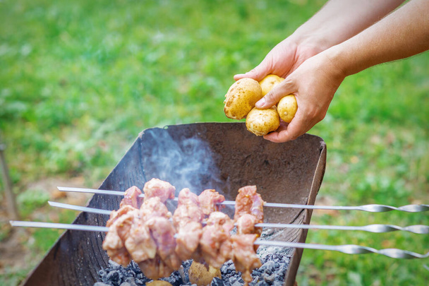 Hands of man prepares barbecue meat with potatoes on skewer by grill on fire outdoors. Concept of lifestyle rustic food preparation - Photo, Image