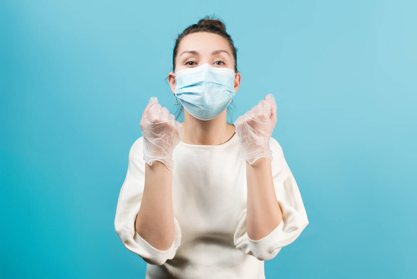 Woman in medical mask and medical gloves clenches fists in mute request. Isolated on a blue background - Fotografie, Obrázek