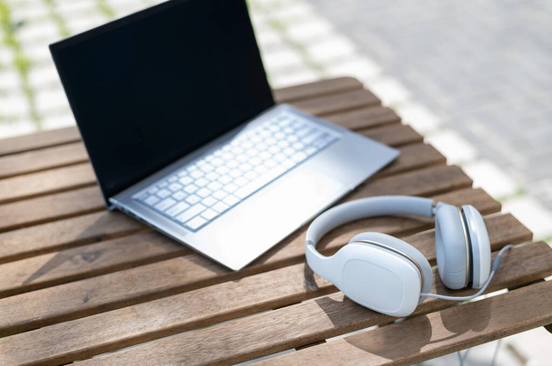 Close-up of a laptop and white big headphones on a wooden table on a summer terrace. No people. Cafe at the outdoors. Freelancer workspace. - Foto, afbeelding