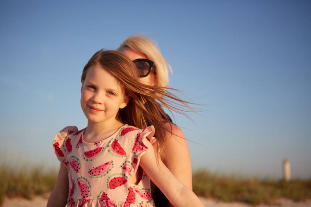 Smiling mother and beautiful daughter having fun on the beach. Portrait of happy woman giving a piggyback ride to cute little girl with copy space. Portrait of kid embracing her mom during summer. - Photo, Image