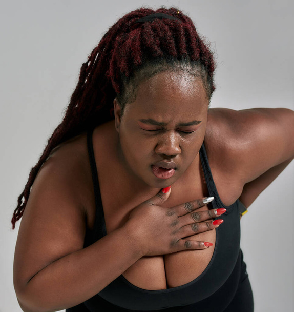 Pain. Close up shot of plump, plus size african american woman in sportswear getting tired, exhausted, feeling pain after workout in studio over grey background - Фото, изображение