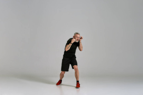 Full length shot of middle aged muscular man in black sportswear practicing punches while exercising in studio over grey background - Photo, Image