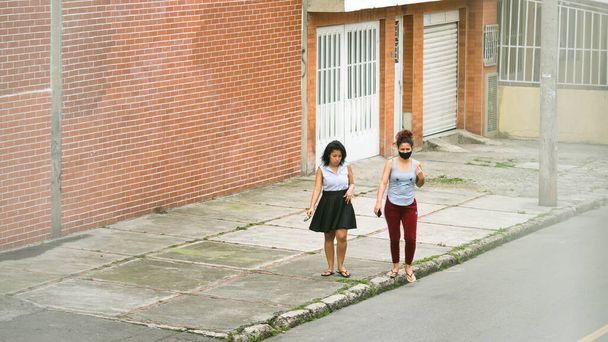Две женщины, молодые и среднего возраста, одна в маске, ходят по улицам Боготы, Колумбия. Избегайте улова и распространения коронавируса. Неофициальная одежда
. - Фото, изображение
