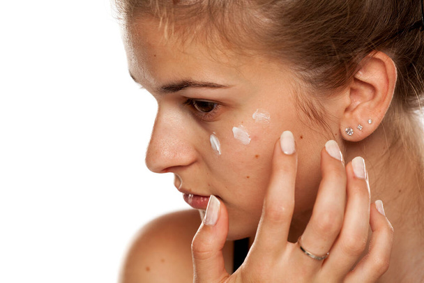 jonge vrouw aanbrengen crème op haar gezicht op witte achtergrond - Foto, afbeelding
