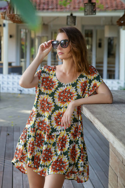 Outdoor portrait of stylish fashionable woman in colorful dress and black sunglasses. Casual summertime hippie boho outfit, spring-summer fashion trend, tropical vacation style. - Photo, Image
