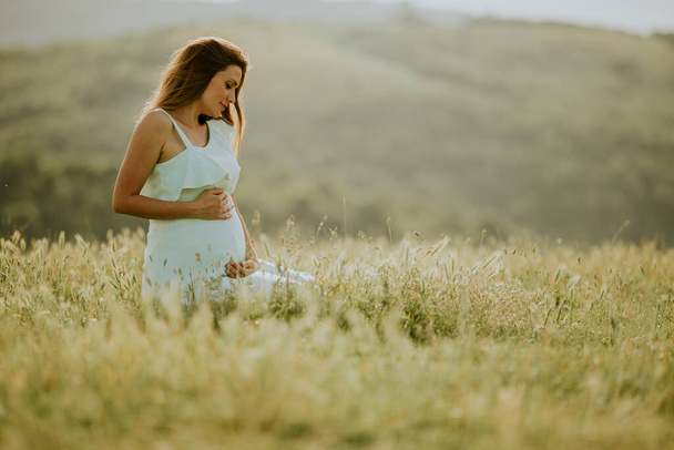 Молода вагітна жінка в білій сукні в літньому полі
 - Фото, зображення