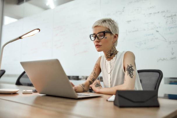 Working on a new project. Young and beautiful tattooed woman with short haircut using laptop while sitting at her working place in the meeting room - 写真・画像