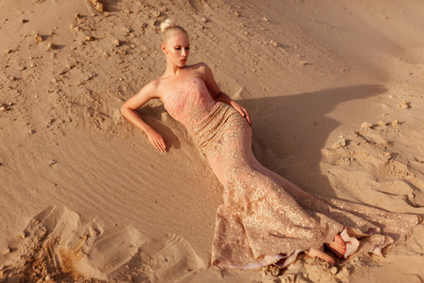 Full length of a blonde woman in expensive gold dress posing in desert, lying on the sand at the sunset. - Fotografie, Obrázek