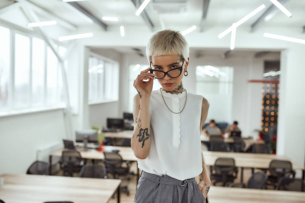 Business lady at work. Confident young and attractive tattooed business woman adjusting her eyeglasses and looking at camera while standing in modern coworking space - Foto, afbeelding