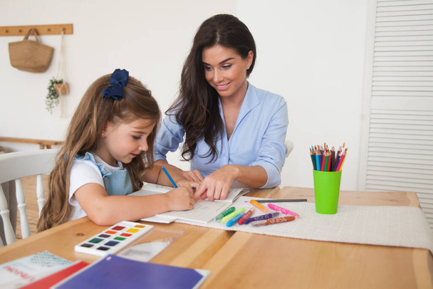 Uśmiechnięta matka i córka przygotowują się do lekcji i rysują przy stole ołówkami i farbami. Rodzic i uczeń przedszkola. Pierwszy dzień jesieni. Dziewczyna z podstawówki, z powrotem do szkoły. - Zdjęcie, obraz