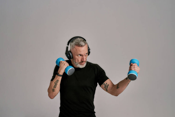 Middle aged muscular man in black t shirt and headphones looking focused holding blue dumbbells, posing in studio over grey background - Photo, Image