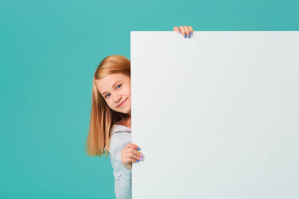 A laughing, happy Caucasian teenage girl looks out from behind an empty whiteboard. Empty space for your text, ads isolated on a turquoise-blue background - Foto, Imagem