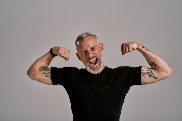 Be more, do more. Angry middle aged muscular man in black t shirt shouting at camera, showing his biceps while posing in studio over grey background - Foto, Imagem