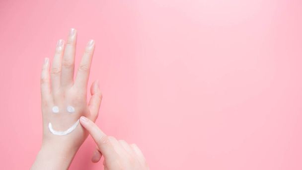 Mooie vrouw handen aanbrengen hydraterende crème op roze achtergrond. Lachende emoji vorm gemaakt van lotion. Zorg voor je lichaam huid. Gezondheidszorg en schoonheidsconcept. Bovenaanzicht. Kopieerruimte. - Foto, afbeelding