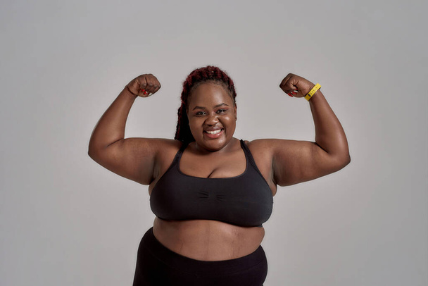 Enjoy your body. Plump, plus size african american woman in sportswear smiling at camera, showing strong arms in studio over grey background. Concept of sport, healthy lifestyle, body positive - Foto, Imagem