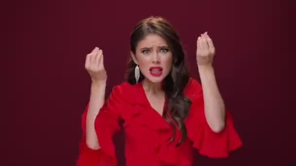 Am emotional angry young woman wearing a red dress swears while raised her hands  isolated over burgundy background - Video